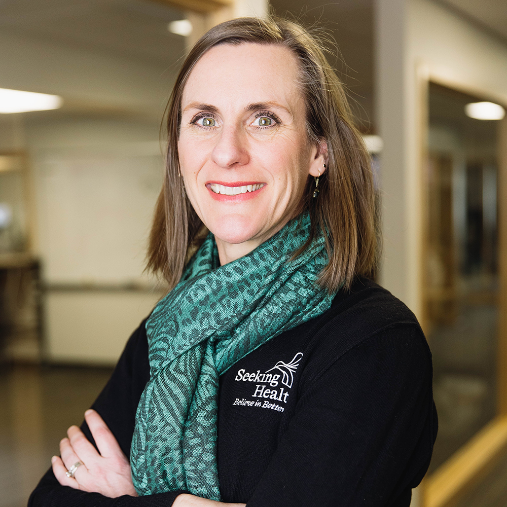 Woman wearing a green scarf with 'Seeking Health' logo in a hallway.