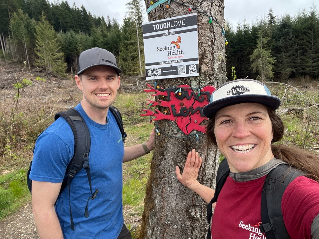 Two people posing next to a tree with a 'Tough Love' sign in a forest setting.