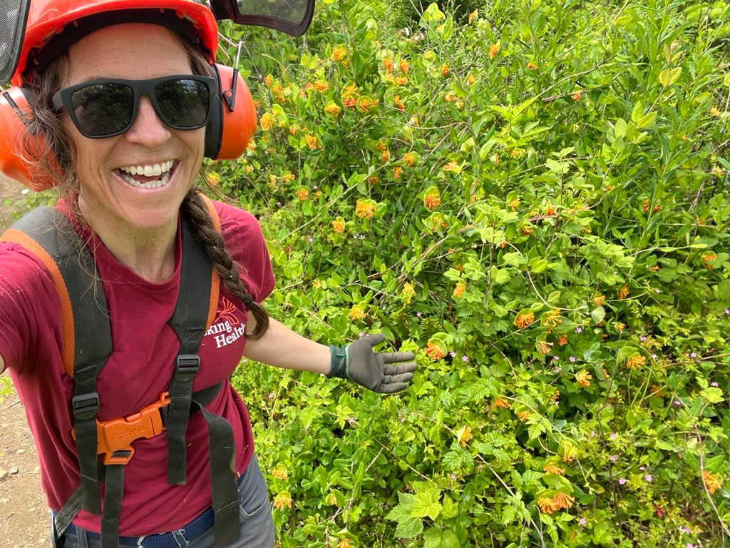 Person in outdoor gear standing next to a bush with green leaves and small orange flowers.
