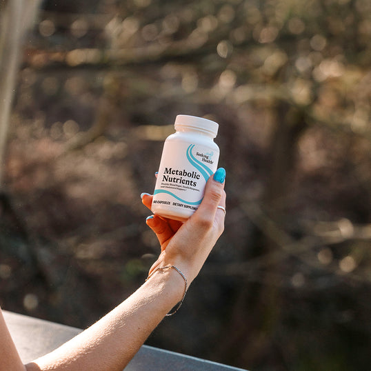 A hand with blue nail polish holds a white bottle of Metabolic Nutrients dietary supplement against a blurred outdoor background of trees.