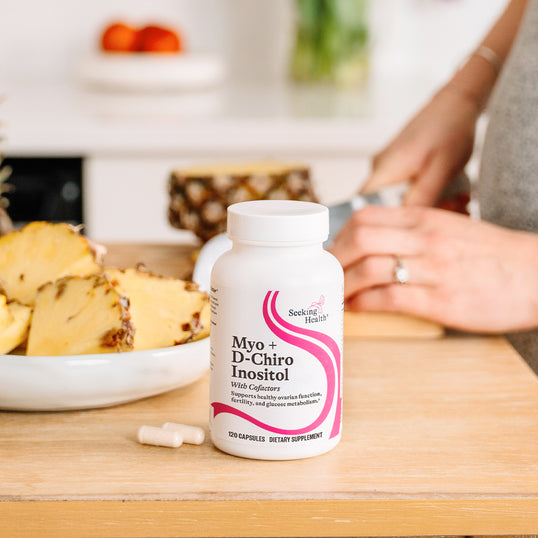 A bottle of Seeking Health Myo + D-Chiro Inositol dietary supplement capsules sits on a wooden cutting board in a kitchen setting, with pineapple slices and a person's hands blurred in the background.