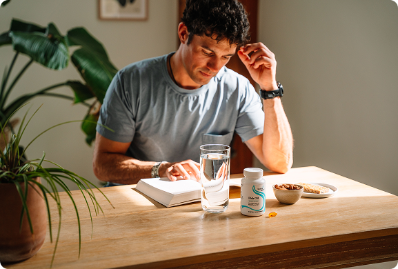 Man sitting at a table with a book, glass of water, and supplement bottle.