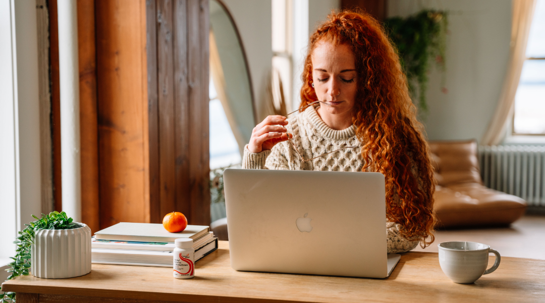 Woman with red hair sitting at a desk using a laptop in a home office setting.