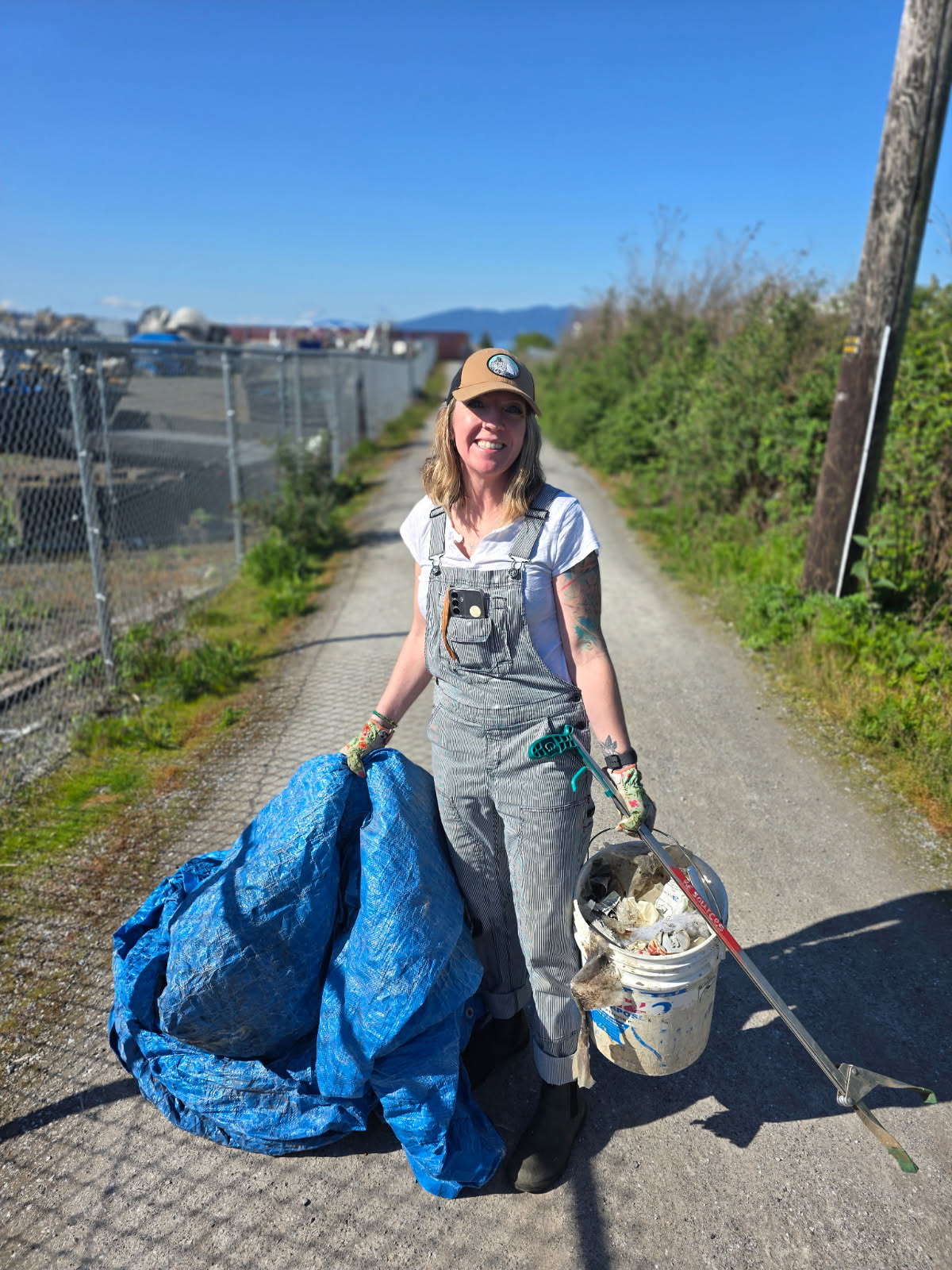 Person holding a blue trash bag and a bucket on a path with greenery and buildings in the background
